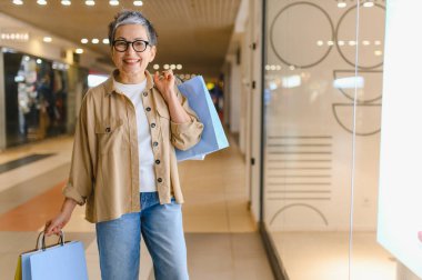 Kıdemli kadın gülümsüyor ve alışveriş torbaları taşıyor, modern bir alışveriş merkezinde başarılı bir tüketim ve yaşam tarzının tadını çıkarıyor.