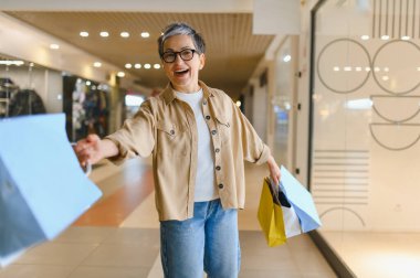Gri saçlı ve gözlüklü neşeli yaşlı kadın gülümsüyor, parlak ışıklı alışveriş merkezi koridorunda renkli alışveriş torbaları taşıyor.