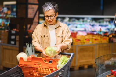 Elinde alışveriş arabası olan yaşlı bir kadın taze marul seçiyor. Dengeli bir diyet için sağlıklı yiyecekler almaya odaklanmış durumda.