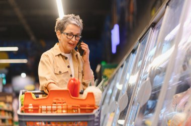 Üst düzey bir kadın akıllı telefonuyla konuşuyor ve markette içi taze ürünlerle dolu bir alışveriş arabasını itiyor.