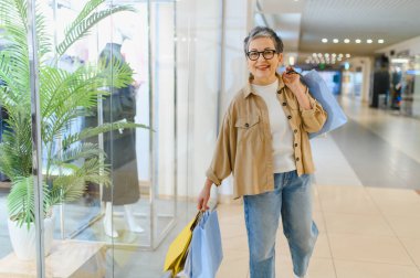 Perakende terapiden hoşlanan mutlu yaşlı bir kadın, elinde alışverişlerle modern bir alışveriş merkezinde yürüyor.