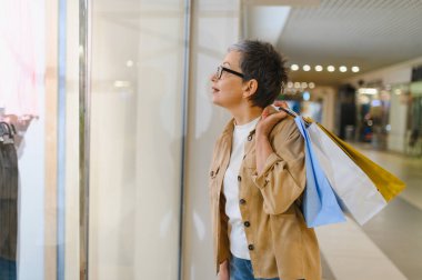 Alışveriş torbaları taşıyan yaşlı bir kadın, modern bir alışveriş merkezinin vitrinine bakıyor. Perakende terapi araştırması