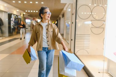 Alışveriş merkezinde yürüyen, vitrinlere bakan, başarılı alışverişlerden sonra renkli alışveriş torbaları taşıyan son sınıf mutlu bir kadın.
