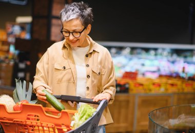 Bakkal koridorunda duran bir kadın taze bir salatalık seçiyor ve alışveriş arabasının içine koyup gülümsüyor.