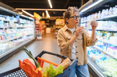 Kadın alışveriş listesi okuyor, markette eşyalar karşılaştırıyor ve yiyecek dolu bir arabayı itiyor. Bütçeye ve perakende satışına odaklan