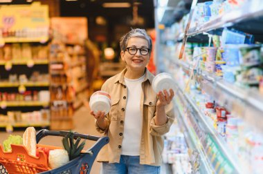 Kıdemli bir kadın gülümsüyor, elinde iki konteynır var. Parlak ışıklı bir süpermarketin sağlıklı gıda bölümünden alışveriş yapıyor.