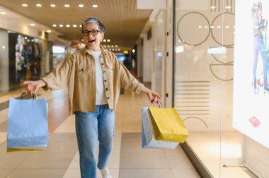 Alışveriş merkezinde elinde renkli çantalarla dolaşan neşeli olgun bir kadın. Alışverişten sonra mutluluk ve heyecan