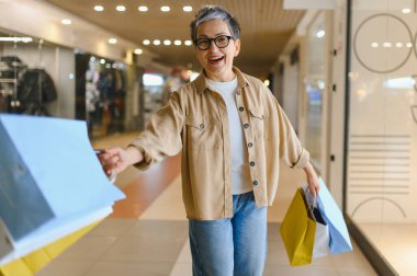 Kadın gülümsüyor, alışveriş merkezinde çantalarla yürüyor, başarılı alışverişleri ve yaşam tarzını kutluyor.