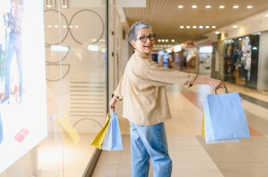 Neşeli yaşlı kadın renkli alışveriş çantaları tutuyor, modern bir alışveriş merkezinde yürüyor, perakende terapisinden ve aktif yaşam tarzından zevk alıyor.