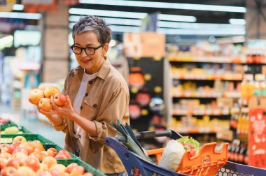 Market koridorundaki vitrinlerden sağlıklı elmaları seçen olgun bir kadın, alışveriş arabasını iterek...