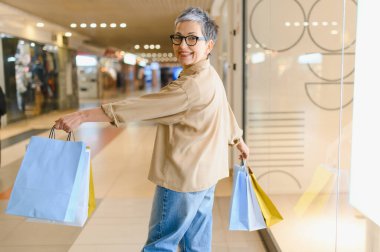 Gri saçlı, gözlüklü, kameraya gülümseyen, alışveriş ve tüketici yaşam tarzının tadını çıkaran olgun bir kadın.