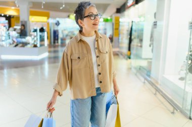 Modern orta yaşlı kadın vitrinlere bakıyor. Parlak bir alışveriş merkezinde alışveriş torbalarıyla mutlu mesut yürüyor.