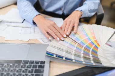 Açık bir paletten bir renk seçen kişinin elleri, mimari planlar ve masadaki bir dizüstü bilgisayarla çalışıyor.