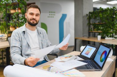 Modern bir ofiste proje üzerinde çalışan genç bir erkek tasarımcı planlar, renk örnekleri ve dizüstü bilgisayarla konseptler planlıyor.
