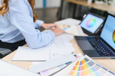 Tasarımcının elleri, dizüstü bilgisayarı, tableti ve mimari planları olan bir masadaki palet kumaşından renkler seçiyor