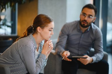 Düşünceli ve endişeli görünen genç bir kadın, bir erkek psikoloğun karşısında oturmuş, akıl sağlığı danışmanlığı sırasında notlar alıyor.