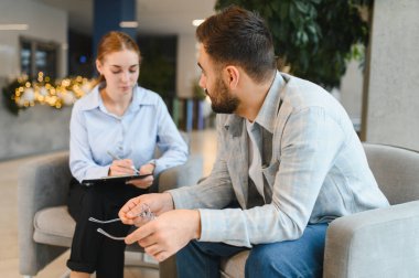 Kadın bir psikologdan danışmanlık alırken elinde gözlük tutan adam profesyonel bir ofiste notlar alıyor.