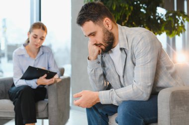 Üzgün adam, modern bir ofiste terapi seansı sırasında bir kadın psikologla ruh sağlığı sorununu tartışıyor.