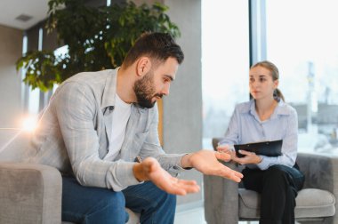 Kanepede oturan erkek hasta modern bir ofisteki bir terapistle sorunlarını konuşurken el kol hareketi yapıyor.