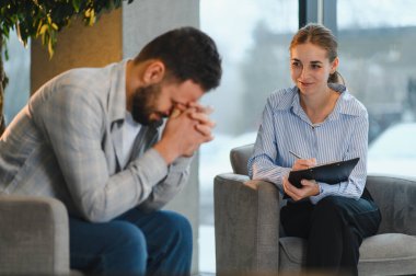Profesyonel bir psikologla terapi seansı sırasında kendini stresli ve depresif hisseden bir adam ofisinde notlar yazıyor.