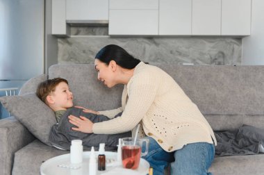 Anne, evdeki kanepede yatan hasta oğluna yardım ve yataklık teklif ediyor. Yanında çeşitli ilaçlar var.