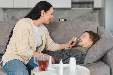 Anne, hasta oğluna boğaz spreyi sıkarak kanepede dinleniyor ve boğaz ağrısı için evde bakım yapıyor.