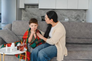 Anne oğluna bakıyor, battaniyeye sarılmış, acı veren boğazını tutuyor, ilaç ve sıcak içecekle sehpanın üzerinde.