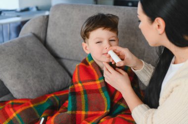 Küçük çocuk kanepede kareli battaniyeye sarılmış soğuk algınlığı veya evde grip tedavisi görürken annesinden burun spreyi alıyor.