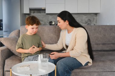 Midesi ağrıyan genç bir çocuk karnını tutarken, annesi ona rahatlık ve ilaç sağlıyor.
