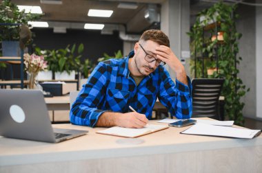 Adam masasında çalışırken yorgun ve baş ağrısından muzdarip. Modern bir ofiste yorgunluk ve baskıyla uğraşıyor.