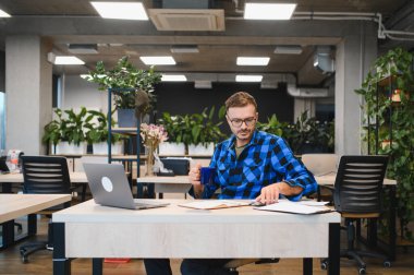 İşadamı birlikte çalıştığı bir masada dizüstü bilgisayar kullanıyor ve sürdürülebilir, fabrika dolu bir çalışma alanında kahve içiyor.