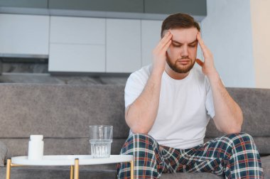 Kanepede başı ağrıyan genç bir adam, migren belirtileri, ilaç tedavisi ve yakınlardaki masada bir bardak su.