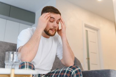 Adam şiddetli baş ağrısı hissediyor, şakaklara dokunuyor ve gözlerini kapıyor. Evde stres, hastalık ve rahatsızlık yaşanıyor.