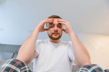 Baş ağrısından dolayı acı çeken genç bir adam parmaklarını şakaklarına bastırıyor. Ev ortamında gözleri kapalı.