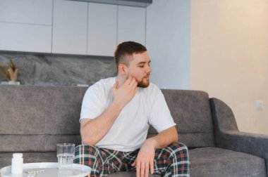 Genç adam pijama giyiyor, kanepede oturuyor ve yanağına dokunuyor, evinde şiddetli bir diş ağrısı çekiyor.