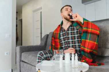 Pijamalı hasta bir adam ve kanepede battaniye. Soğuk algınlığı veya grip için ilaç alıyor. Evde huzur buluyor.