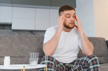 Şiddetli baş ağrısı ya da migren geçiren genç bir adam, masada ilaç ve su ile bir koltukta oturuyor, rahatlamaya ihtiyacı var.