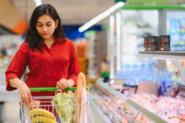 Hintli bir kadın taze ürünlerle dolu bir alışveriş arabasını itiyor, markette sağlıklı bir yemek için eşyalar seçiyor.