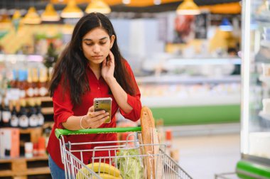 Marketteki alışveriş arabalarını iten, ürünlerini karşılaştıran ve alışveriş listesini bir süpermarkette kontrol eden genç bir kadın.