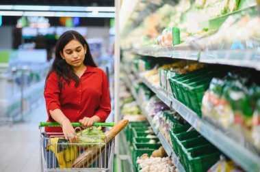 Hintli bir kadın marketin rafından sağlıklı yiyecek, taze sebze ve ekmek seçiyor, alışveriş arabasını itiyor.