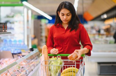 Genç bir kadın alışveriş arabasını itiyor, süpermarket raflarından ürünler seçiyor, taze yiyecek ve günlük ihtiyaçlar alıyor.