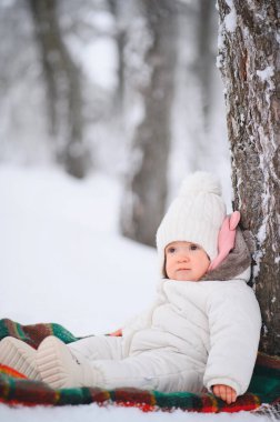 Bebek, karlı bir ormanda, ağaç gövdesinin yanında, renkli bir battaniyede oturuyor. Beyaz kış kıyafetleri içinde.