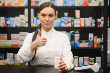 Eczacı gülümsüyor, baş parmak işareti yapıyor, elinde ilaç şişesi tutuyor, eczanede kaliteli sağlık hizmeti veriyor.