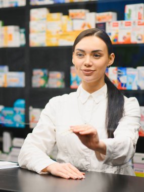 Genç bayan eczacı müşteriye hizmet ediyor. Elinde haplarla kasada duruyor.
