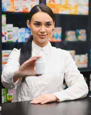 Tezgahın arkasındaki modern eczanede kadın eczacı müşteriye gülümsüyor ve beyaz ilaç şişesini sunuyor.