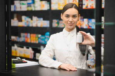 Tezgahta duran eczacı, elinde beyaz bir ilaç şişesiyle eczane raflarının bulanık arka planına karşı duruyor.