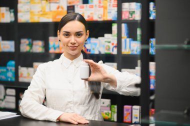 Eczacı gülümsüyor, elinde beyaz bir ilaç şişesiyle ilaç ürünleriyle dolu rafların bulanık arka planına yaslanıyor.