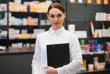 Beyaz üniformalı genç bir kadın eczacı elinde bir pano, çağdaş bir eczanede çalışıyor.
