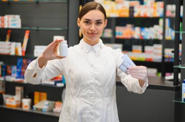 Modern bir eczanede çalışan, elinde ilaç şişesi ve su toplayan beyaz önlüklü, gülümseyen bir kadın eczacı. Sağlık hizmetleri yardımı sağlanıyor