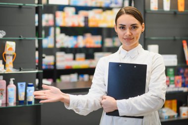 Beyaz önlüklü bir kadın eczacı elinde bir panoyla eczanede durup ilaç ve ürünlerle dolu rafları işaret ediyor.